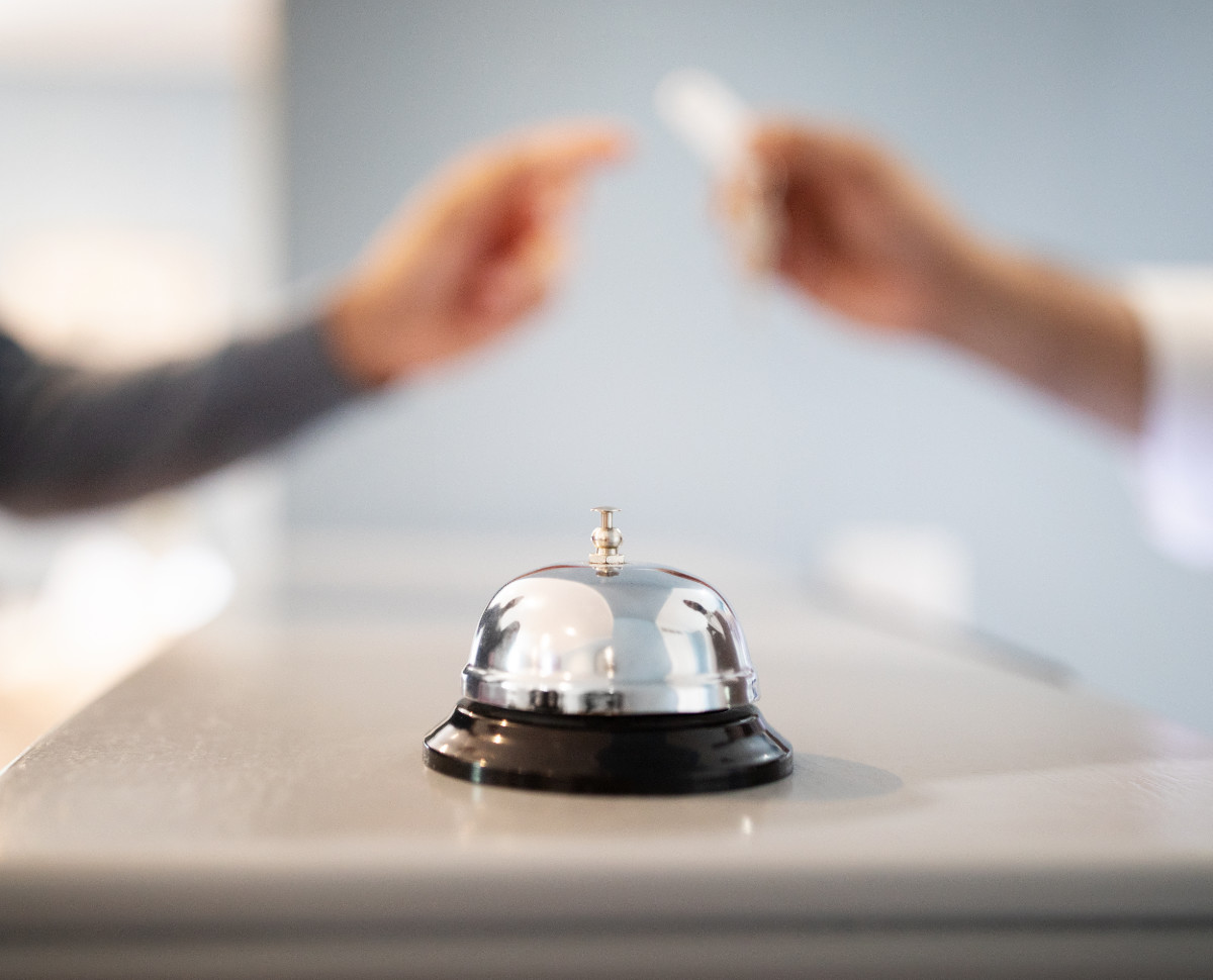 Call Bell Standing On Reception Counter At Hotel Lobby While Receptionist Giving Key To Tourist Man, Selective Focus, Closeup. Guest Checks In At Accommodation, Takes Room Keys From Hostel Staff